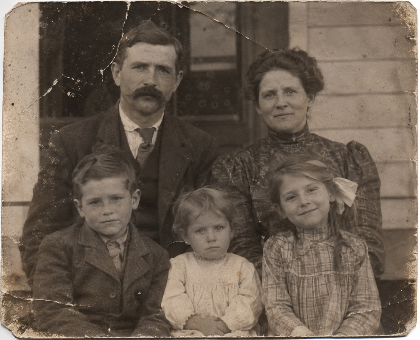 Early 1900s sepia family portrait on a porch: two adults and three children, with cracks across the print